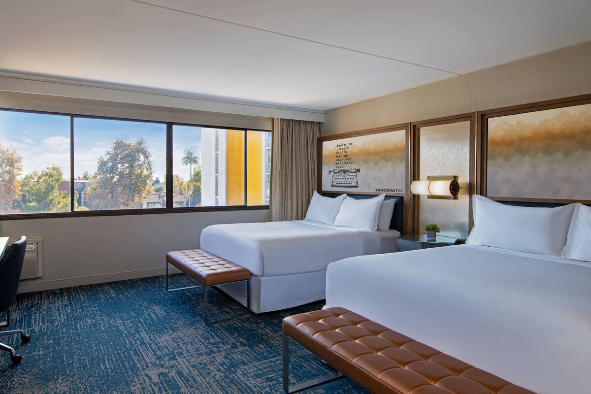 A hotel room with two beds, a desk, and a window with a view of trees and buildings.