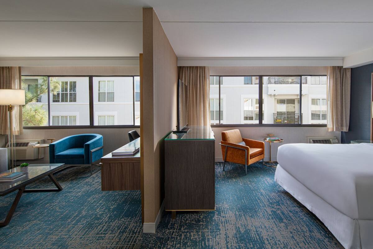 A hotel room with a large bed, two armchairs, a desk, and a small table. There are large windows with curtains, a blue carpet, and a view of buildings outside.