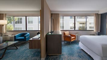 A hotel room with a large bed, two armchairs, a desk, and a small table. There are large windows with curtains, a blue carpet, and a view of buildings outside.