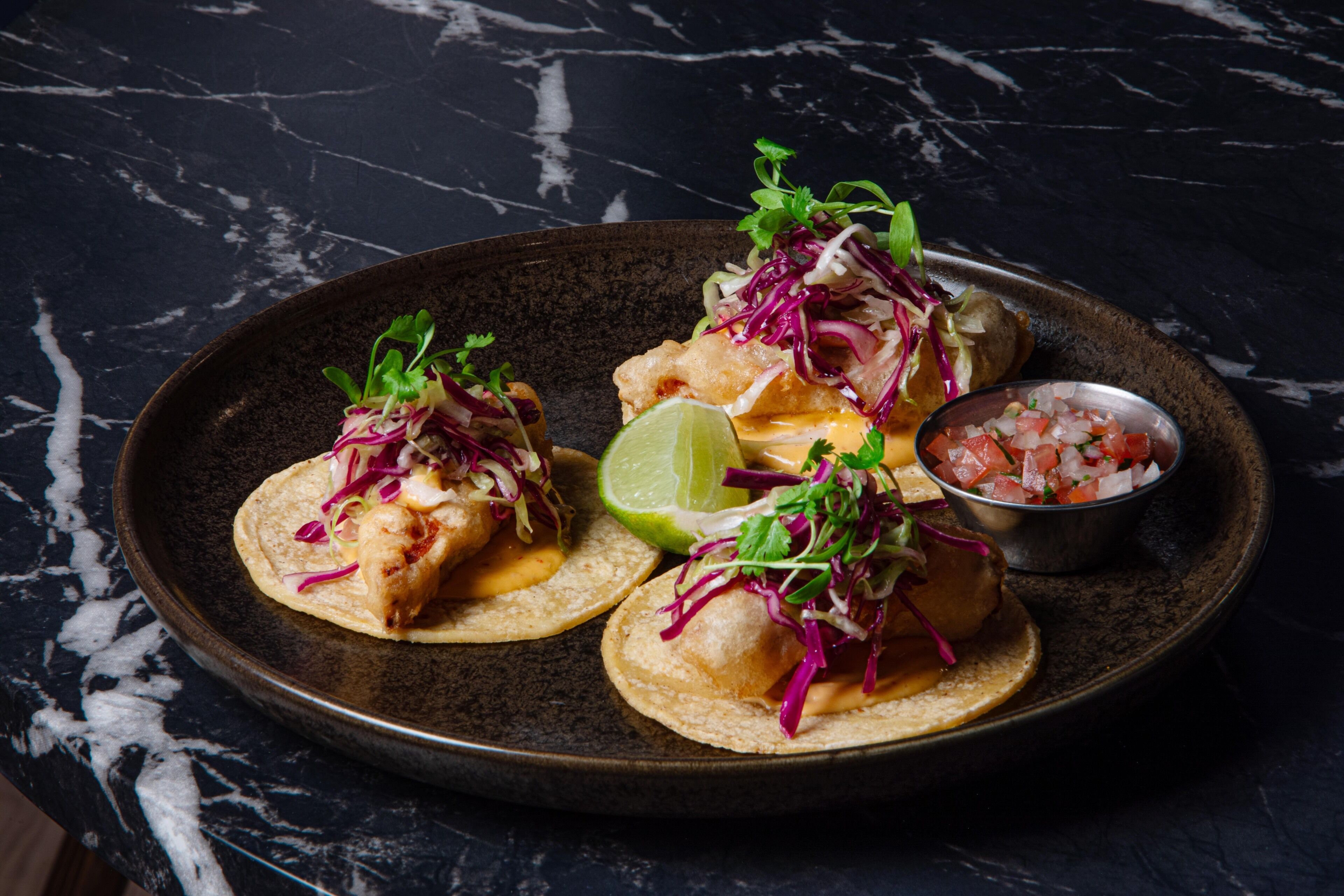 Een bord taco's met limoen en een klein schaaltje salsa op een donkere marmeren ondergrond.