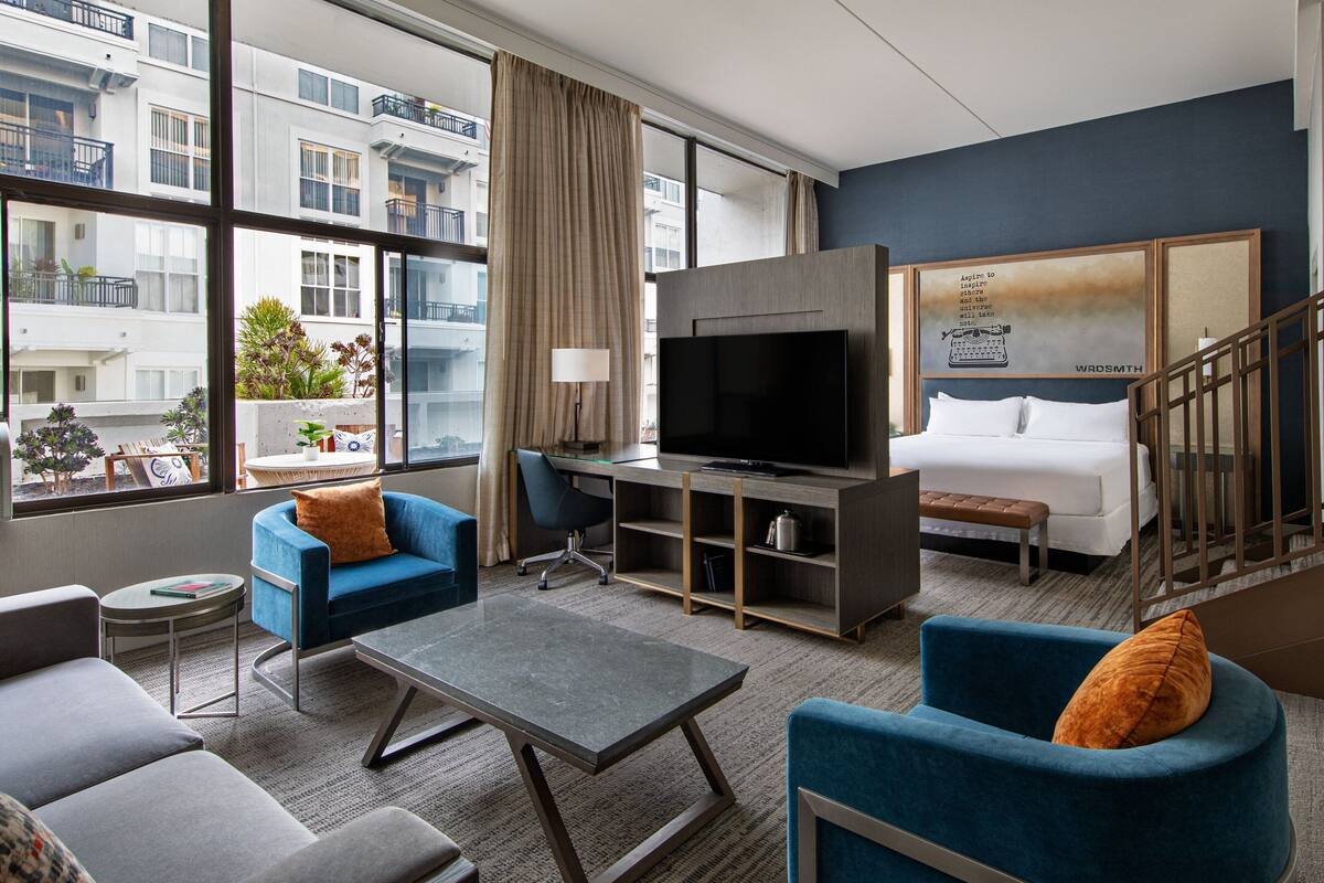 A modern hotel room with a large TV, comfortable seating, a desk, and a view of the city.