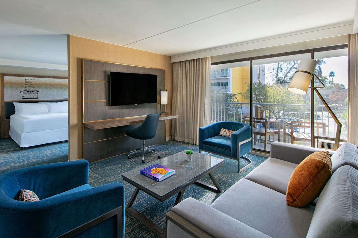 A hotel room with a large bed, a sofa, two armchairs, a coffee table, a TV mounted on the wall, and a balcony with a view of the city.