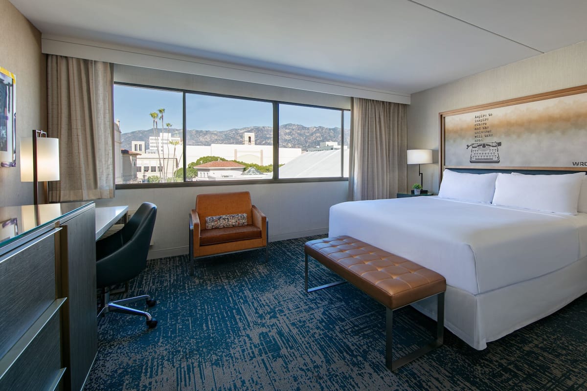 A hotel room with a large bed, a desk, a chair, and a view of the cityscape.