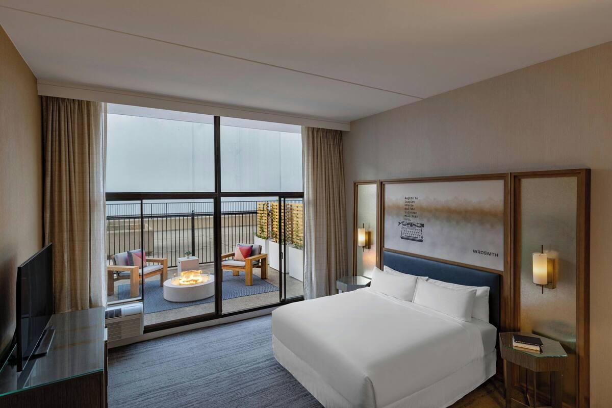 A hotel room with a large bed, a TV, a small table, and a balcony with seating and a fire feature.
