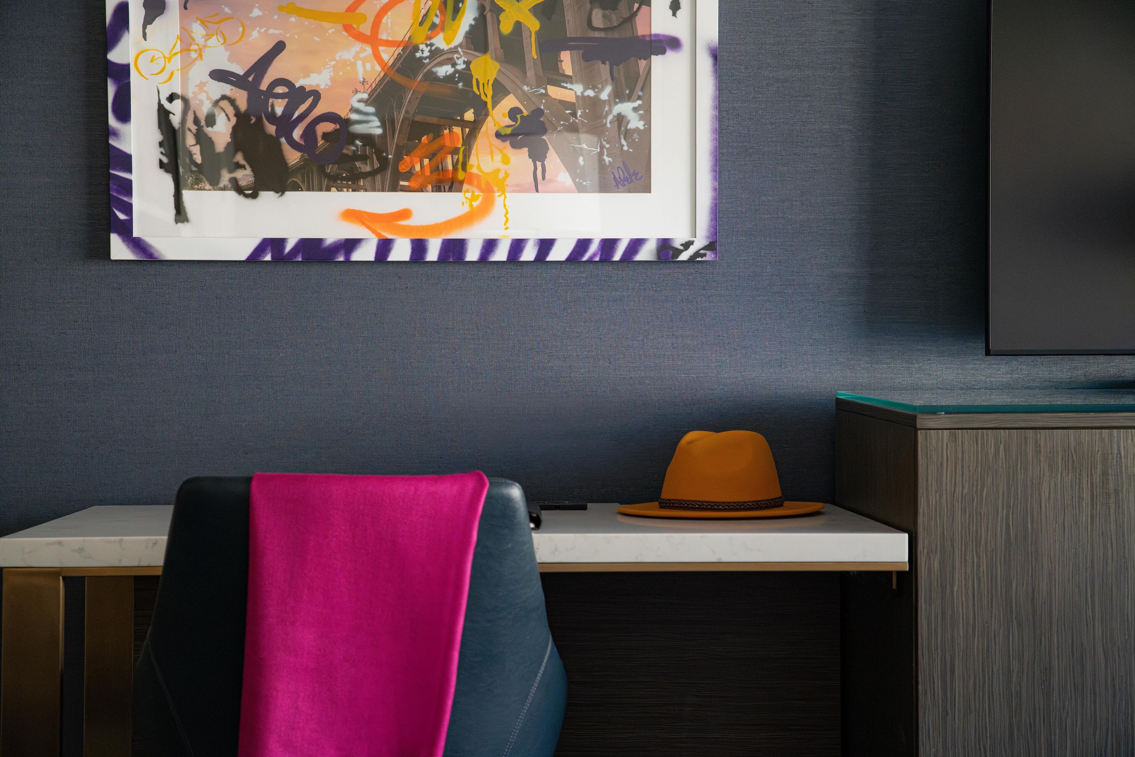 A room with a desk, a chair with a pink and blue cover, a hat on the desk, and a large abstract painting on the wall.