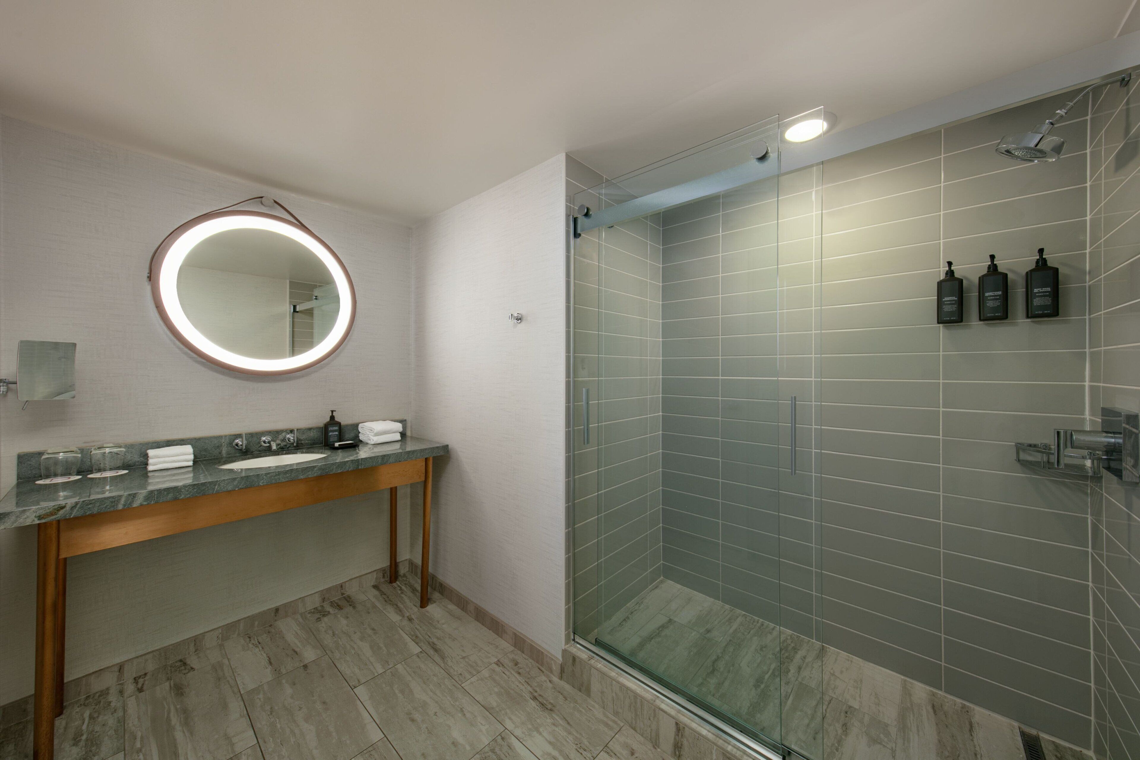 Une salle de bain moderne avec une douche à parois de verre, un miroir rond avec éclairage intégré et un comptoir en marbre.