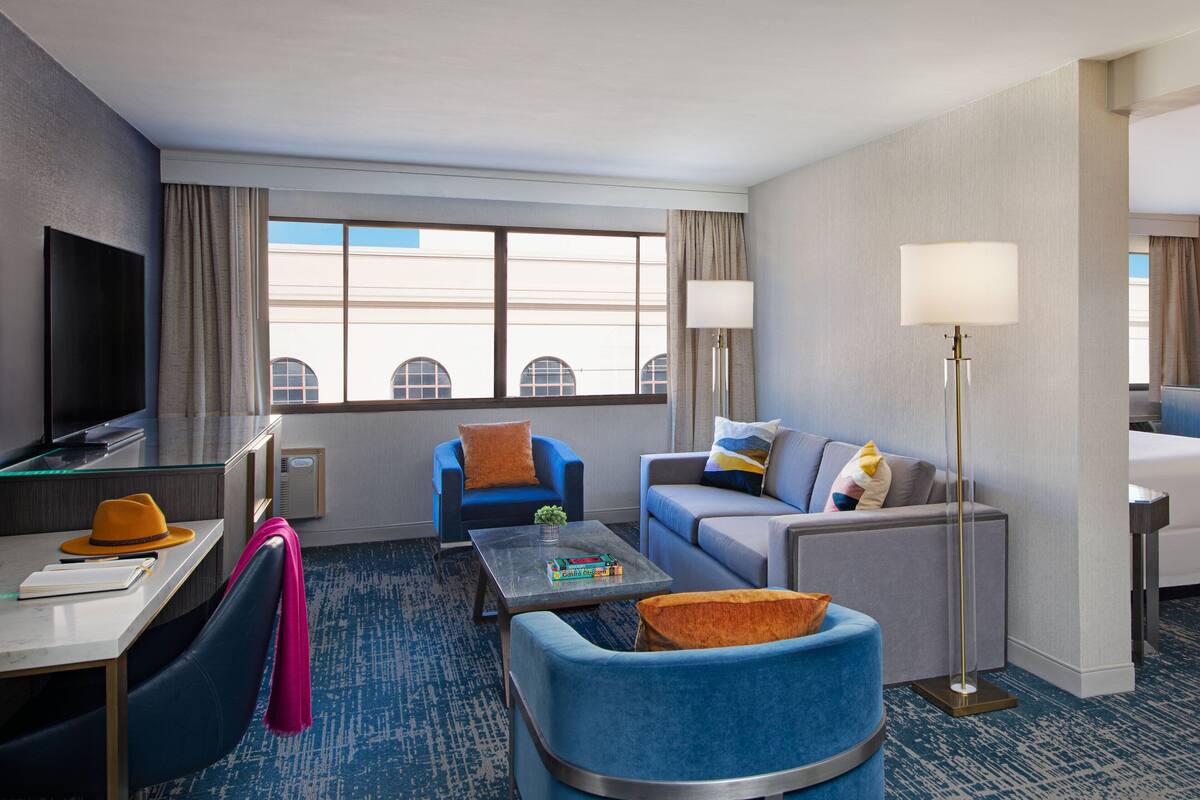 A hotel room with a flat-screen TV, a desk with a chair, a sofa, a coffee table, and a floor lamp.