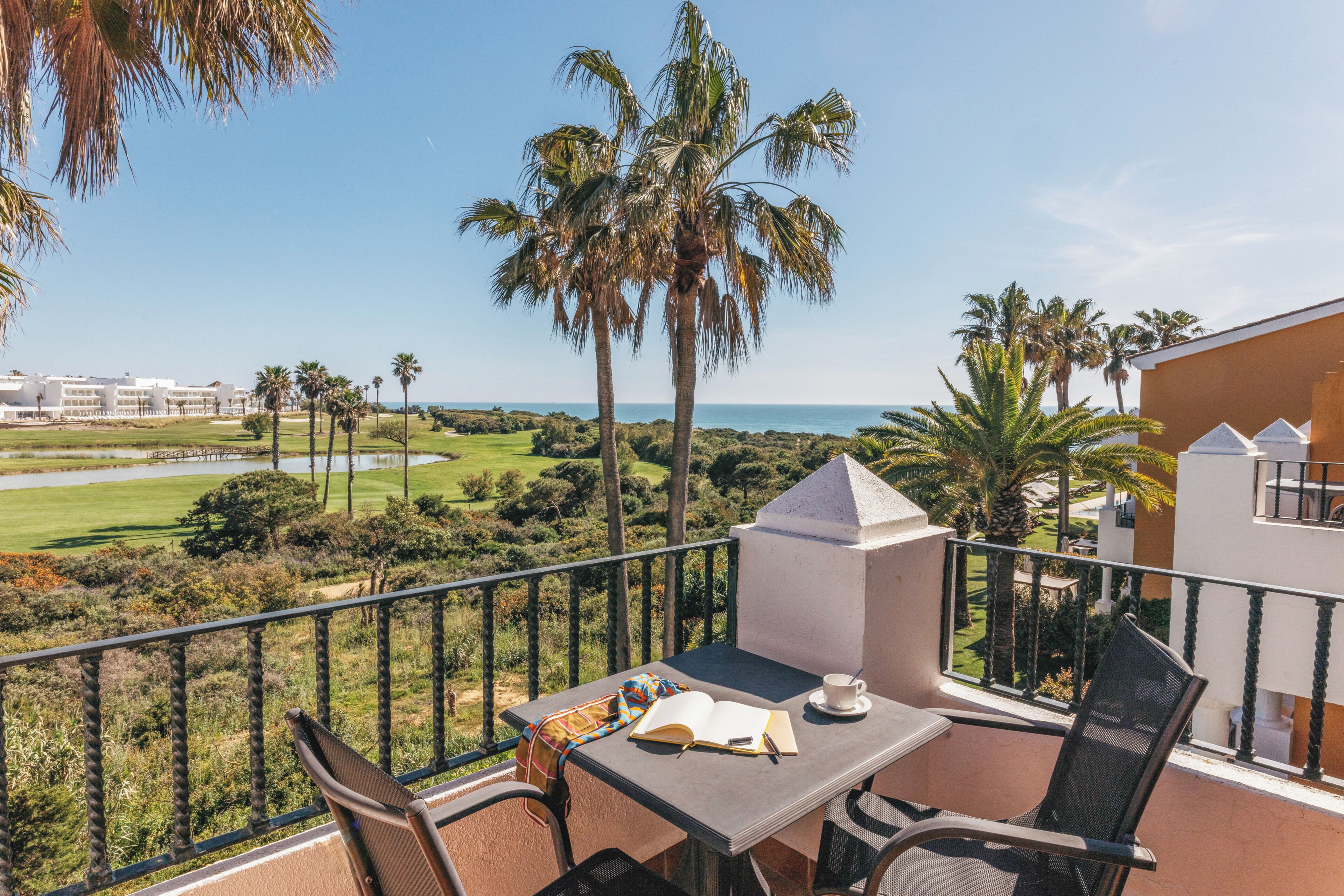 Un balcón con una mesa, sillas y un libro, con vistas a un campo de golf y palmeras.