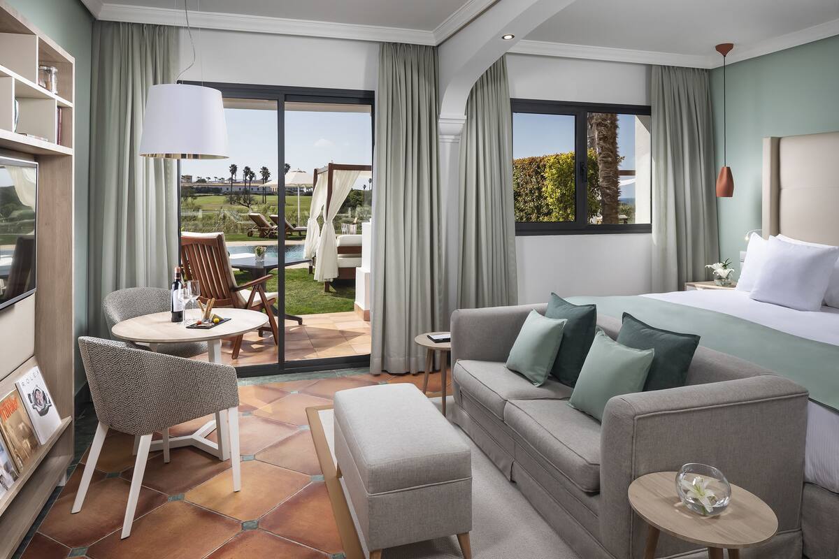 A modern hotel room with a large bed, a sofa, a dining area with a table and chairs, and a view of the outdoor garden and pool.