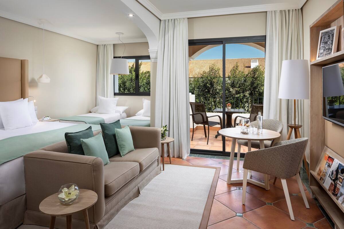 A hotel room with a bed, a sofa, a small table with a lamp, a chair, and a view of the outside.