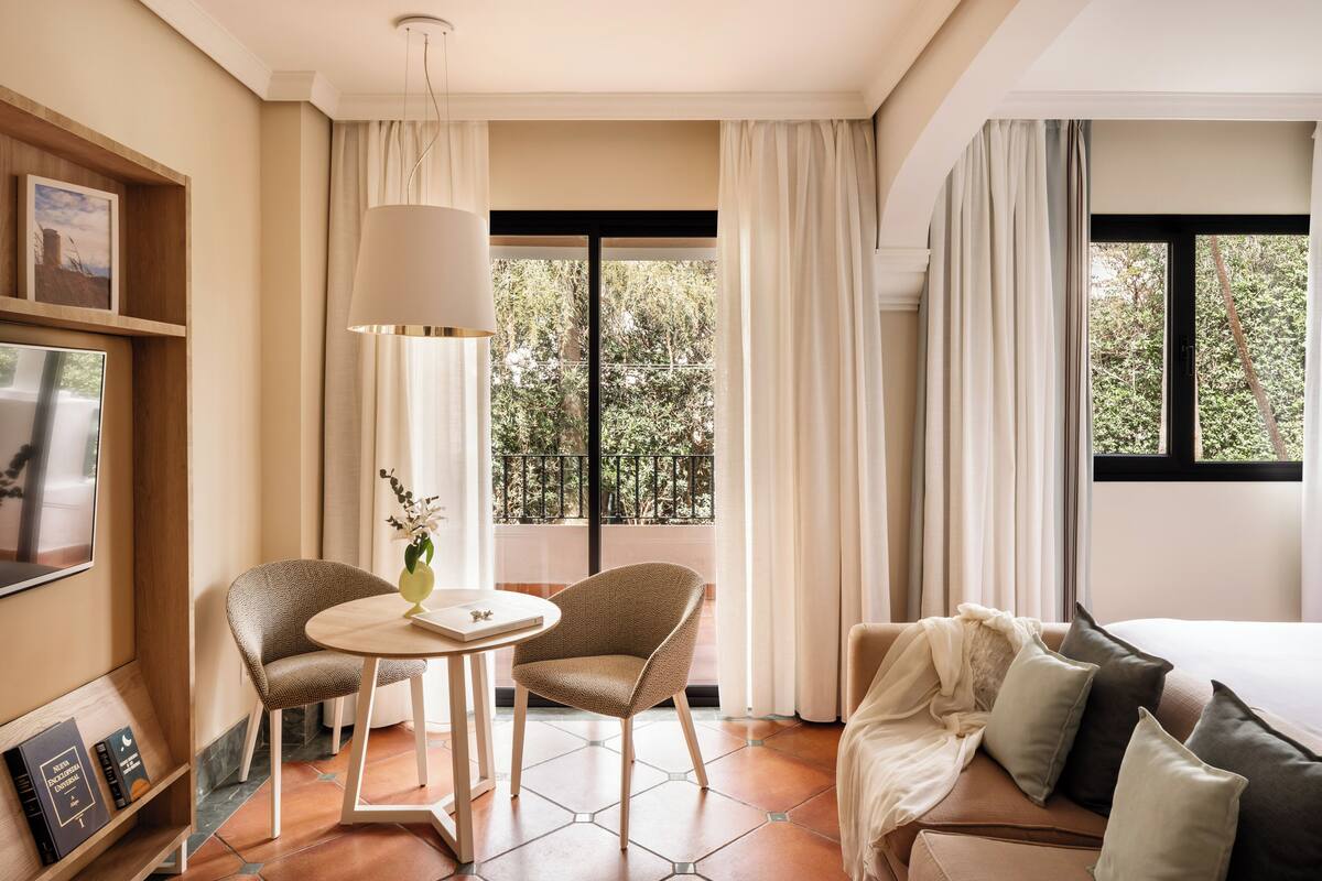 A hotel room with a bed, two armchairs, a small table, and a TV on the left wall.