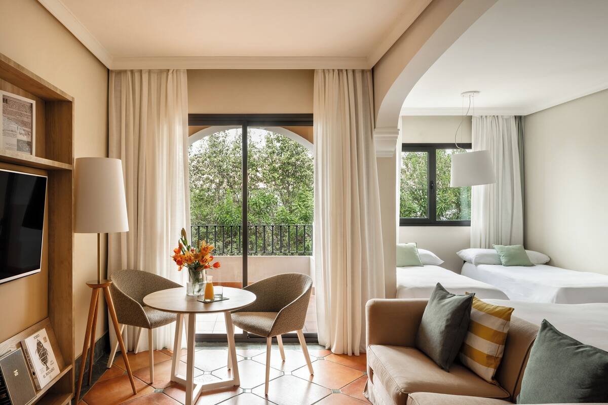 A hotel room with a balcony, a dining area with a table and chairs, a sofa, and a bed.