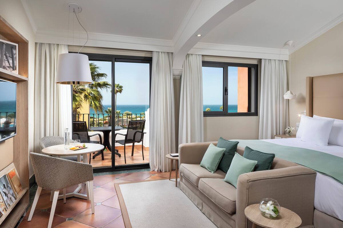 A hotel room with a balcony, a bed, a sofa, a dining table, and chairs.