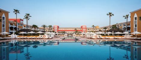 A hotel pool area with a central reflecting pool, lounge chairs, umbrellas, and palm trees.
