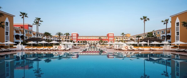 A hotel pool area with a central reflecting pool, lounge chairs, umbrellas, and palm trees.
