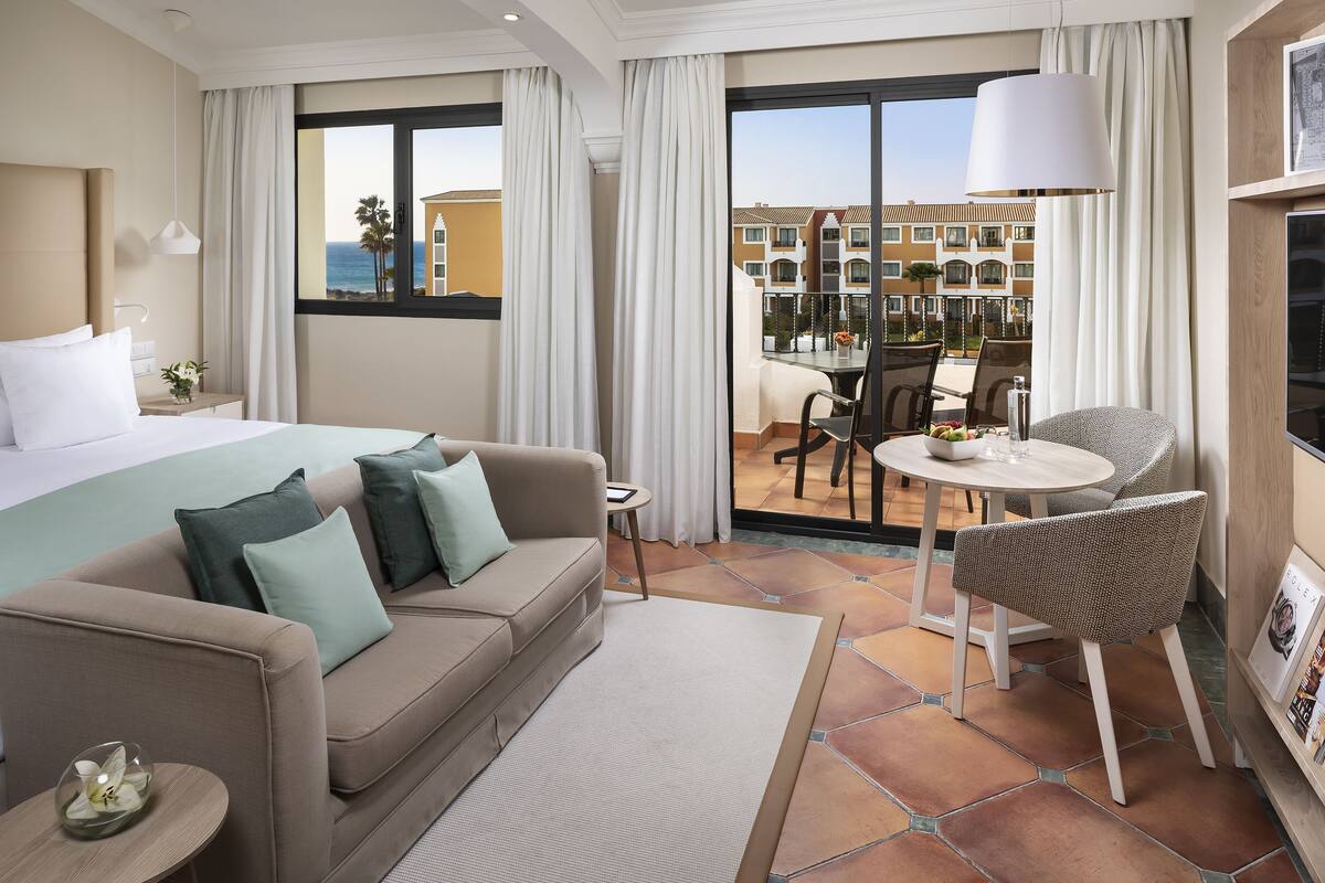 A hotel room with a bed, a sofa, a dining table, and a chair. There is a view of the outside balcony and buildings.
