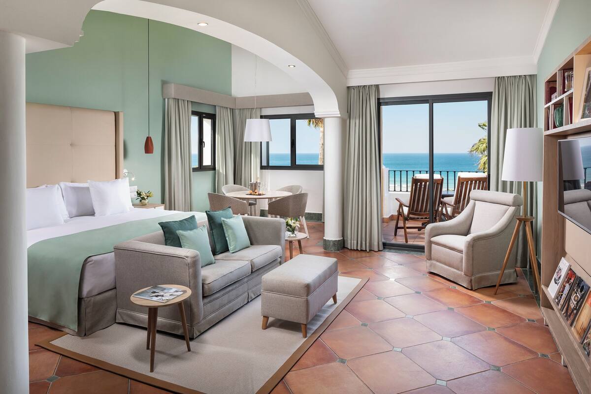 A hotel room with a bed, a sofa, a small table, a chair, and a balcony with a view of the sea.