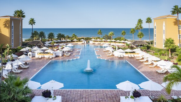A resort pool with a central fountain, surrounded by sun loungers and umbrellas, with a view of the sea and palm trees. - Palacio de Sancti Petri, a Gran Meliá - The Leading Hotels of the World (Chiclana de la Frontera)
