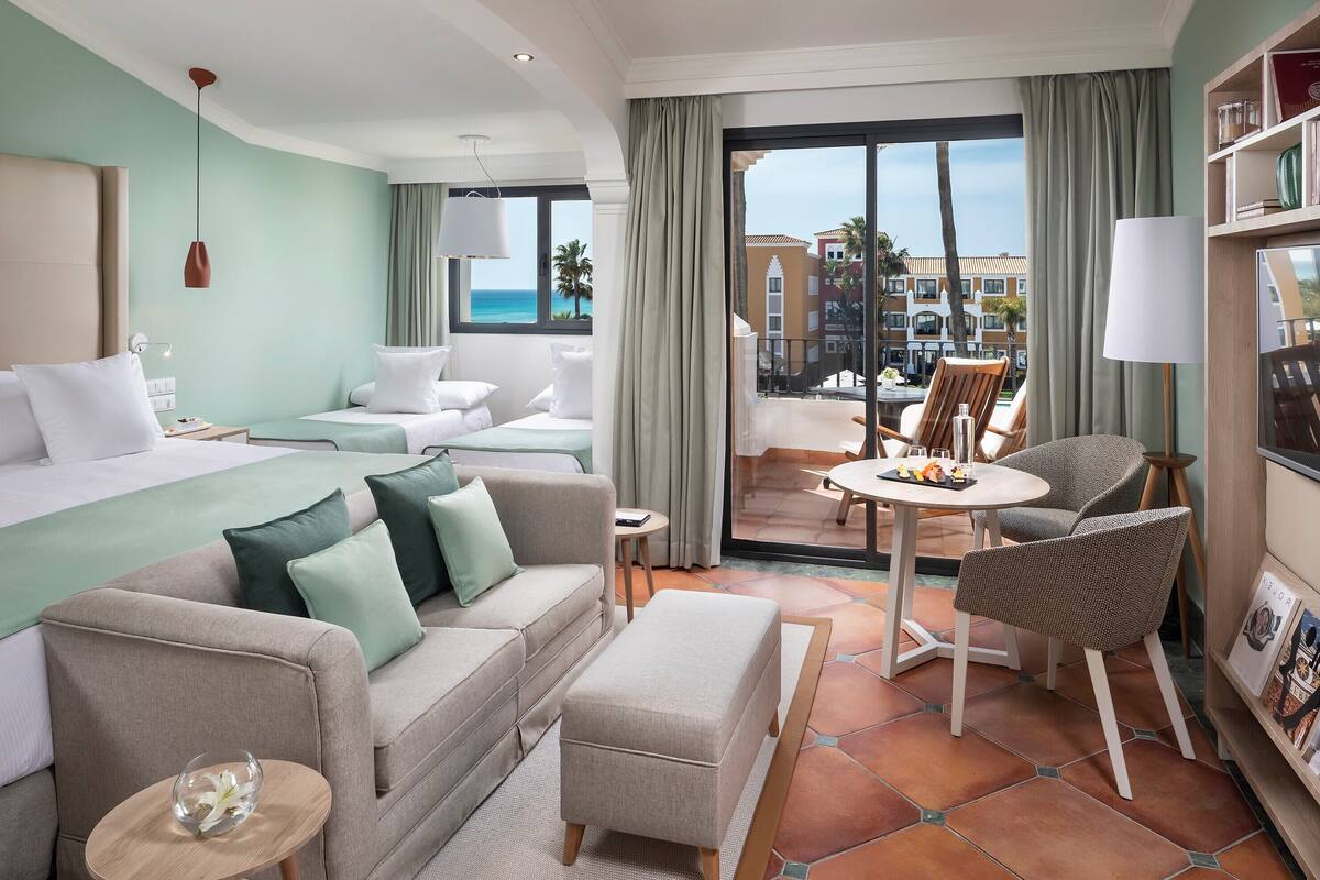 A hotel room with a bed, a sofa, a small table, and a chair. There is a balcony with a table and chairs outside.