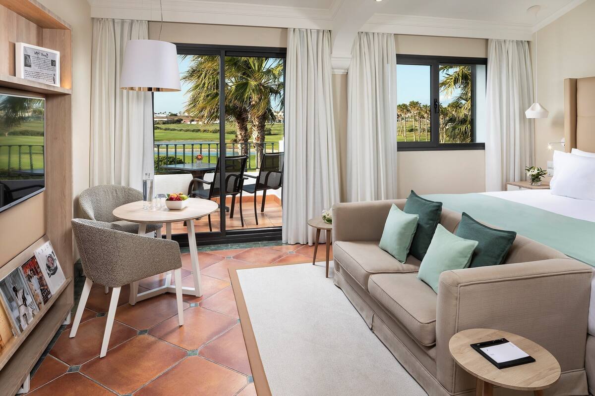 A hotel room with a bed, a sofa, a chair, a small table, and a view of a golf course.