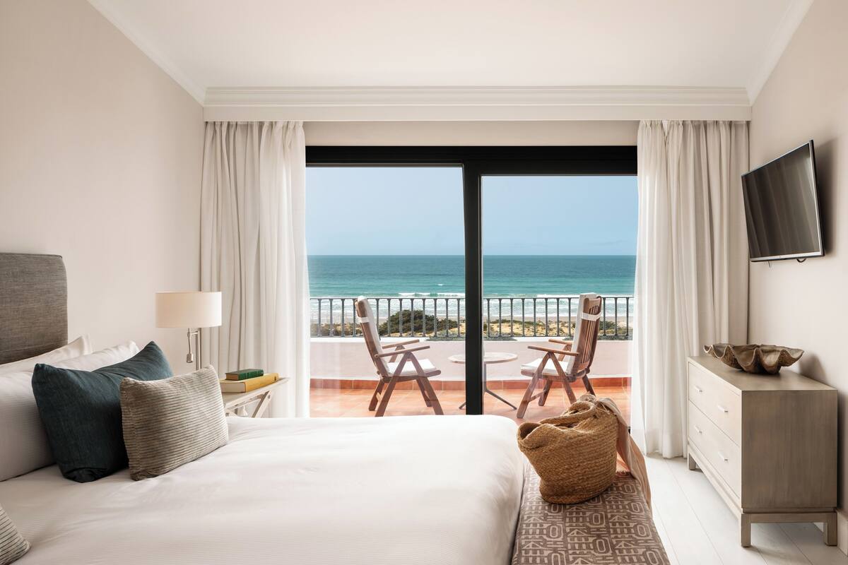 A hotel room with a bed, a TV, a balcony with a view of the sea, and a dining area with chairs and a table.