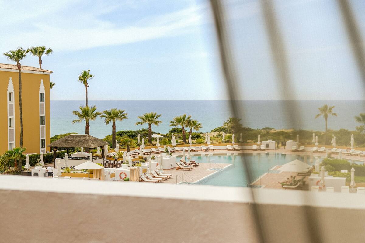 A resort pool area with lounge chairs and umbrellas, a building with a yellow facade, and palm trees.