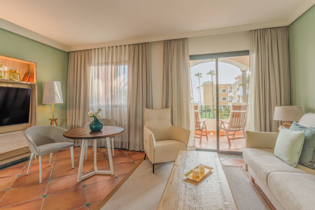A hotel room with a sofa, armchair, and a table with a vase. There is a TV on the left and a balcony with chairs.