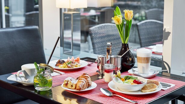 A breakfast setting with croissants, a fruit bowl, a coffee cup, and a vase with flowers on a table. - Lindner Hotel Cologne Am Dom, part of JdV by Hyatt (Cologne)