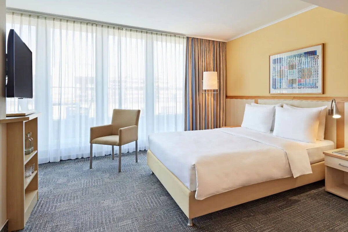 A hotel room with a bed, a chair, a desk, a lamp, and a television.