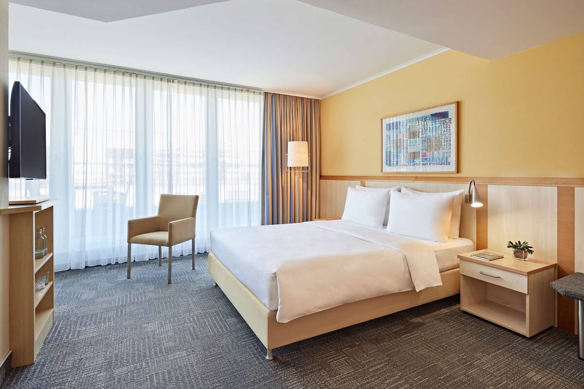 Una habitación de hotel con cama, una silla, escritorio, lámpara y televisión.
