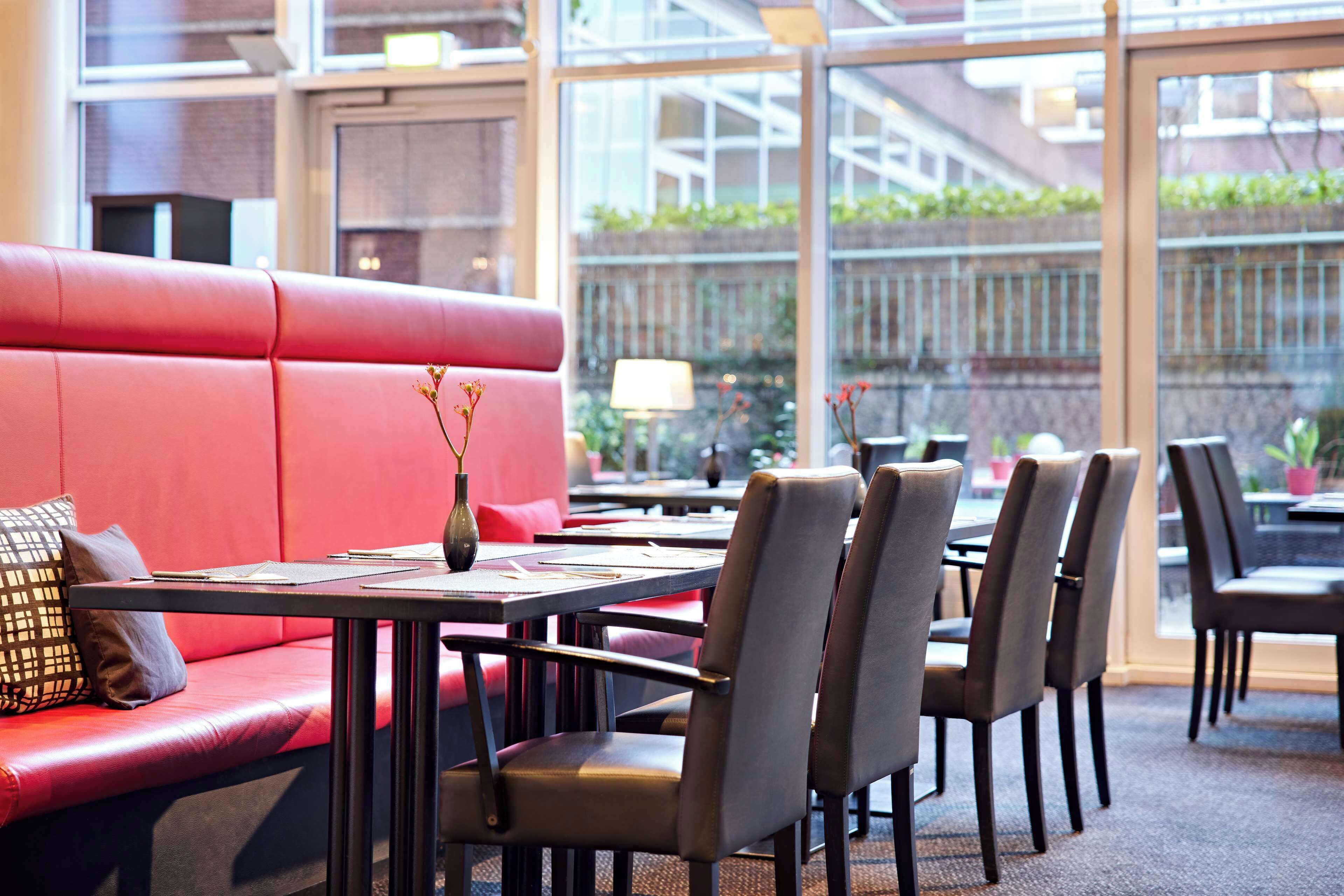 A red cushioned seating area with tables and chairs, a vase with flowers, and a view of a brick wall and greenery outside.