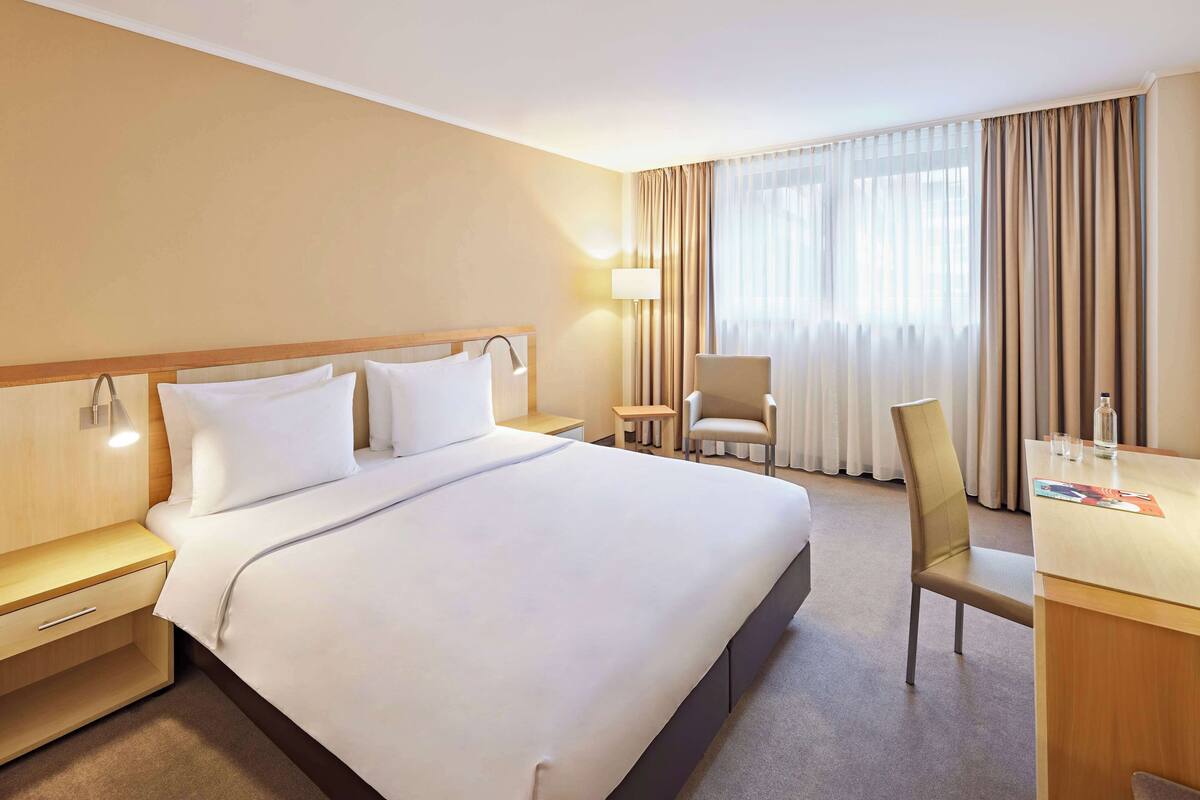 Habitación de hotel con una cama grande, un escritorio, dos sillas, una lámpara y vistas al exterior a través de cortinas translúcidas.