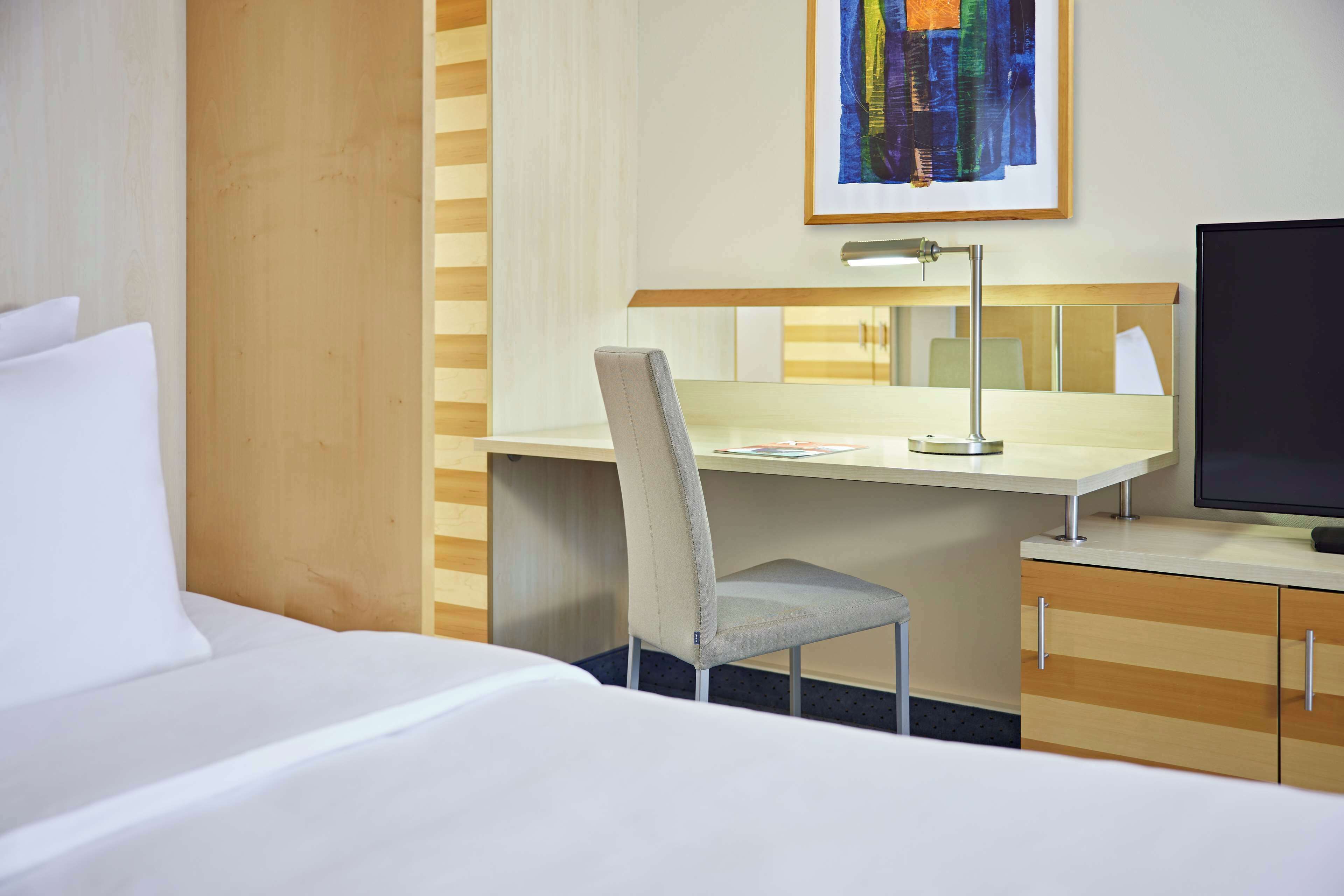 A hotel room with a bed, a desk with a chair, a desk lamp, a television, and a painting on the wall.