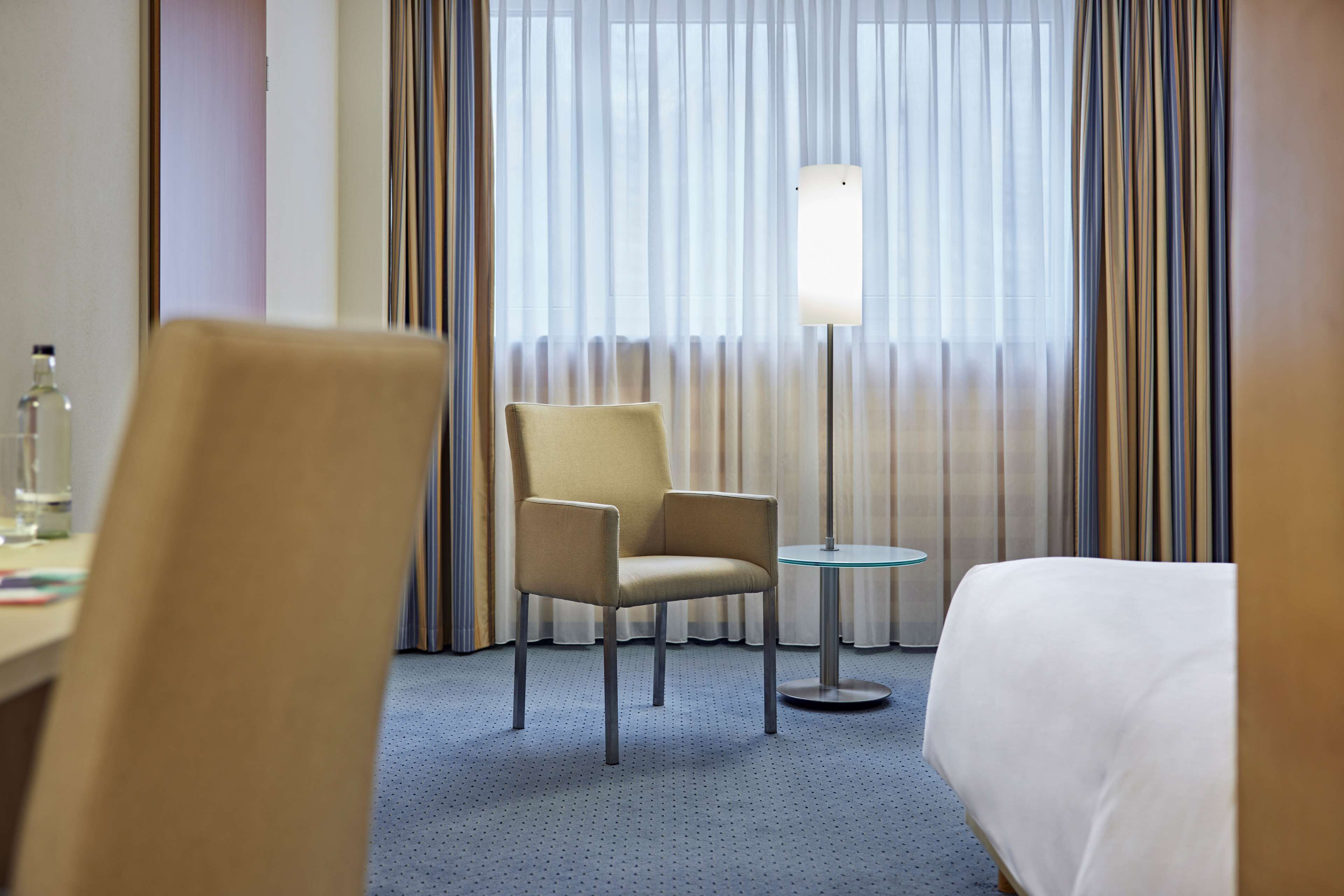 A hotel room with a bed, a chair, a small table, and a lamp.
