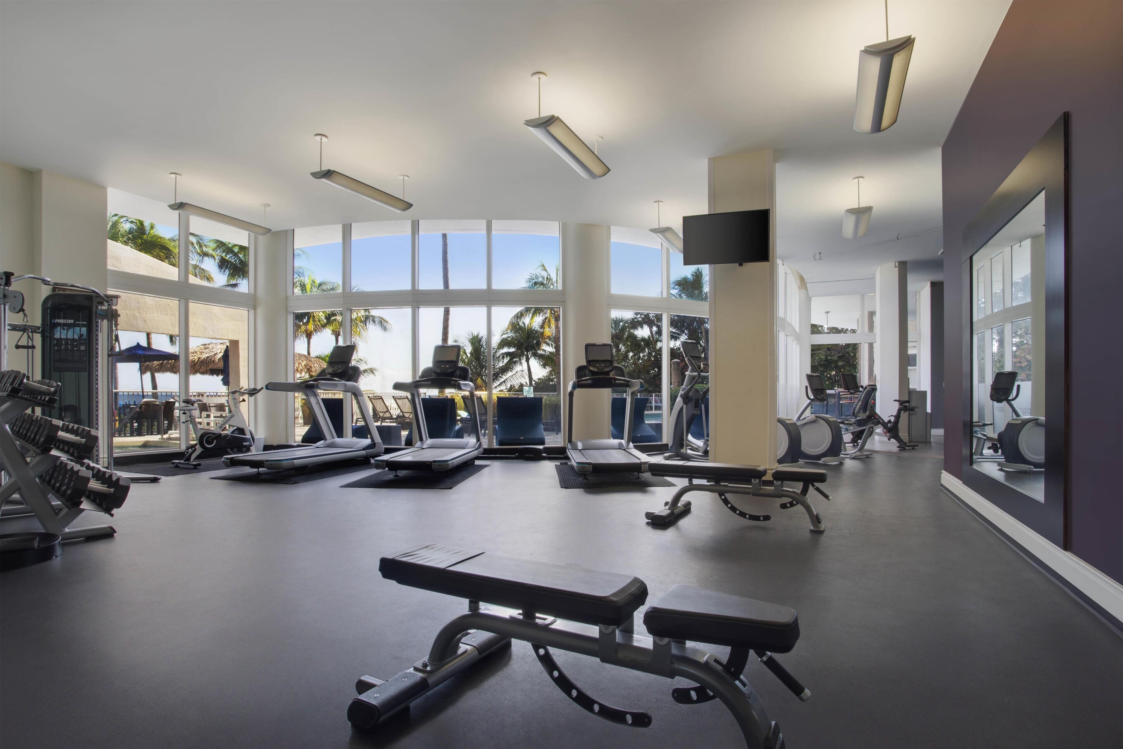 Une salle de sport équipée de divers appareils d’exercice, dotée de grandes fenêtres et offrant une vue sur des palmiers.