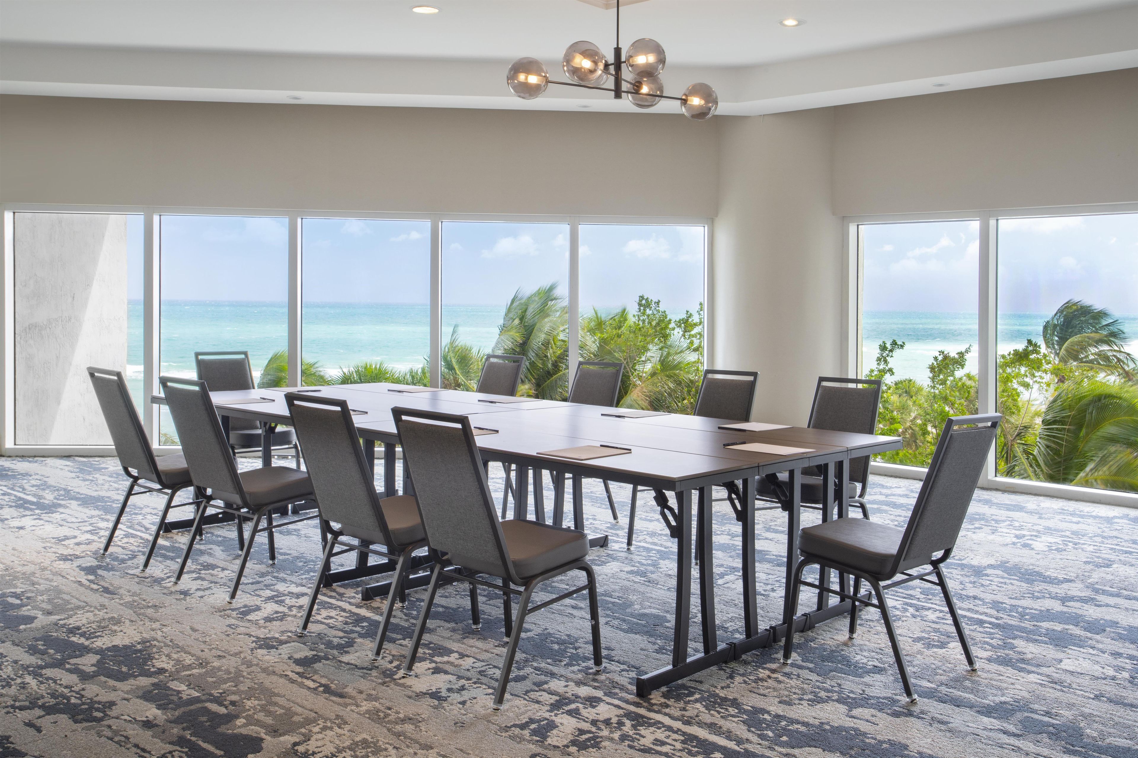 A conference room with a large table and chairs, large windows with a view of the ocean, and a modern chandelier.