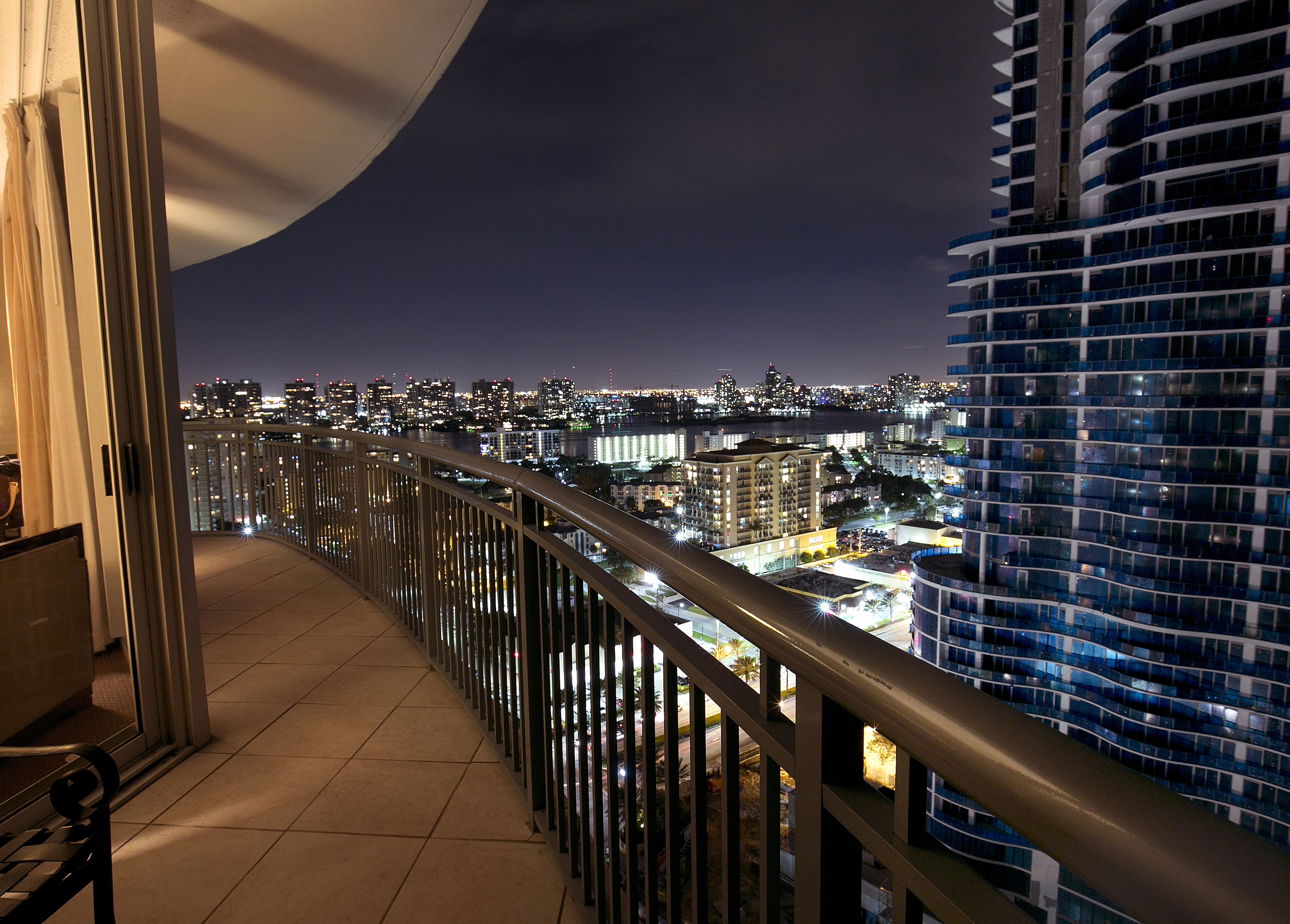A balcony with a view of a cityscape at night, featuring high-rise buildings and a river.
