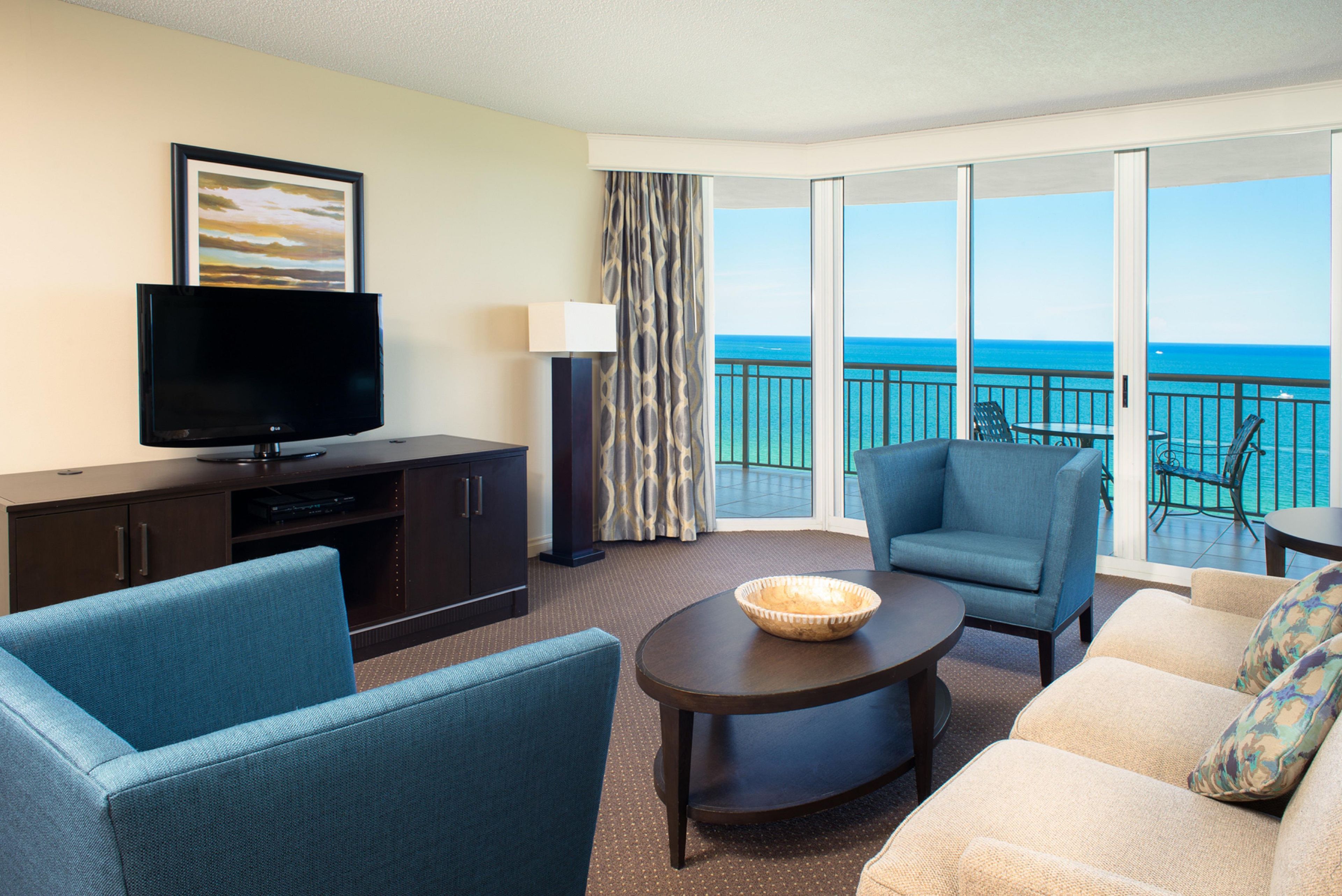 A hotel room with a flat-screen TV, a sofa, a chair, a small table, and a view of the ocean through large windows.