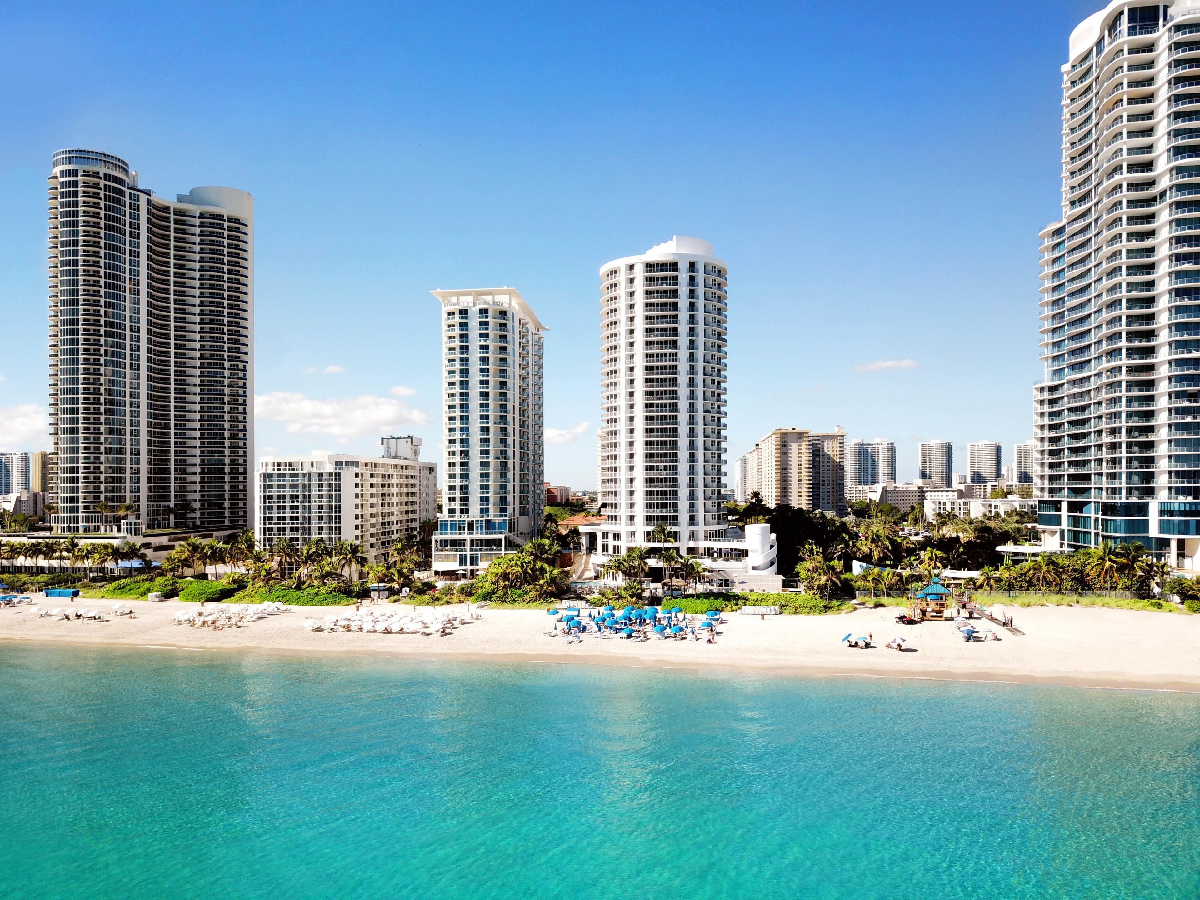 Un hotel frente a la playa con varios edificios altos, una playa de arena y aguas turquesas cristalinas.