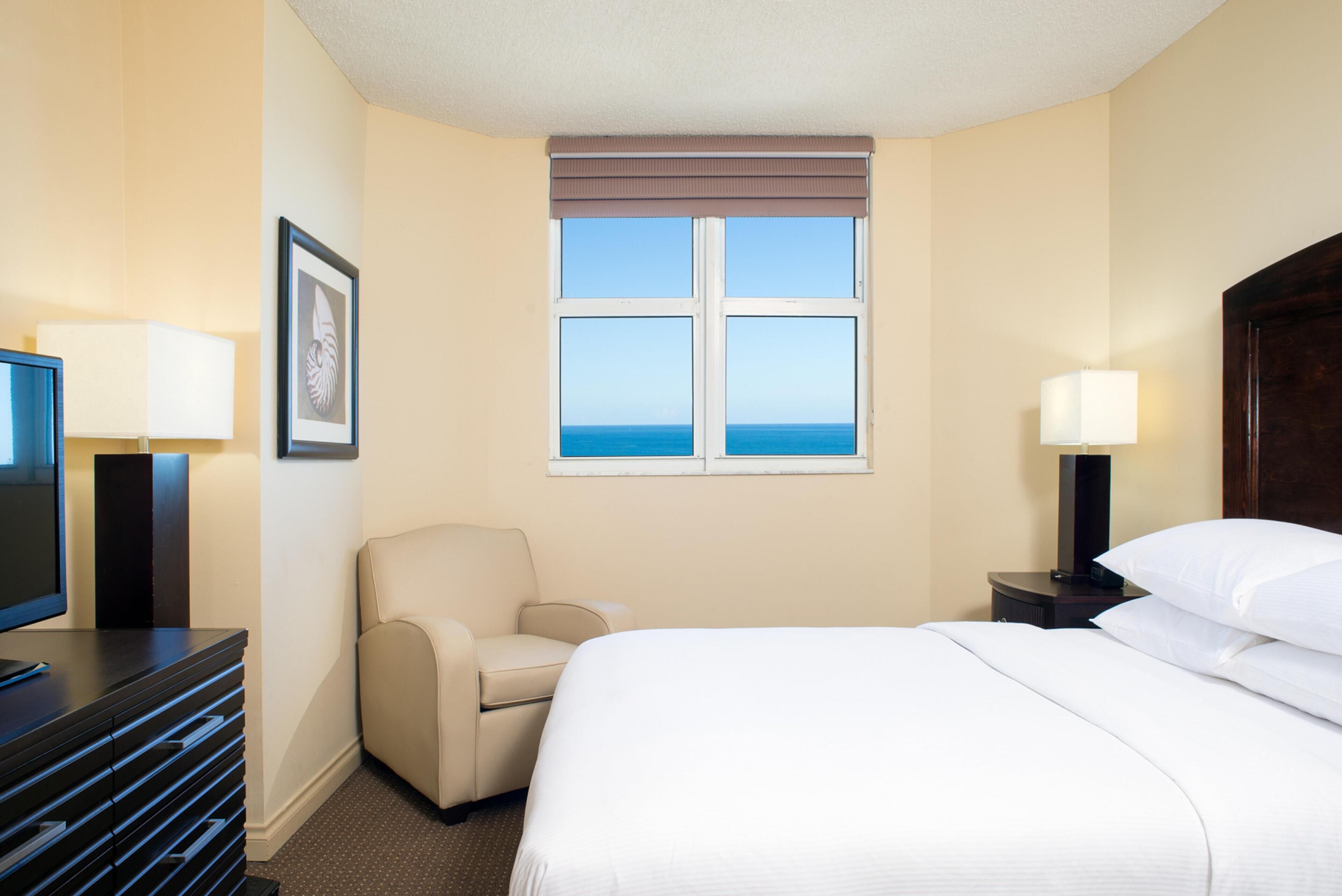 Una habitación de hotel con una cama, una silla, un escritorio con lámpara, un televisor y una ventana con vistas al mar.