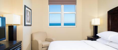 Una habitación de hotel con una cama, una silla, un escritorio con lámpara, un televisor y una ventana con vistas al mar.