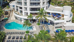 A multi-story hotel with a pool, sun loungers, and palm trees.
