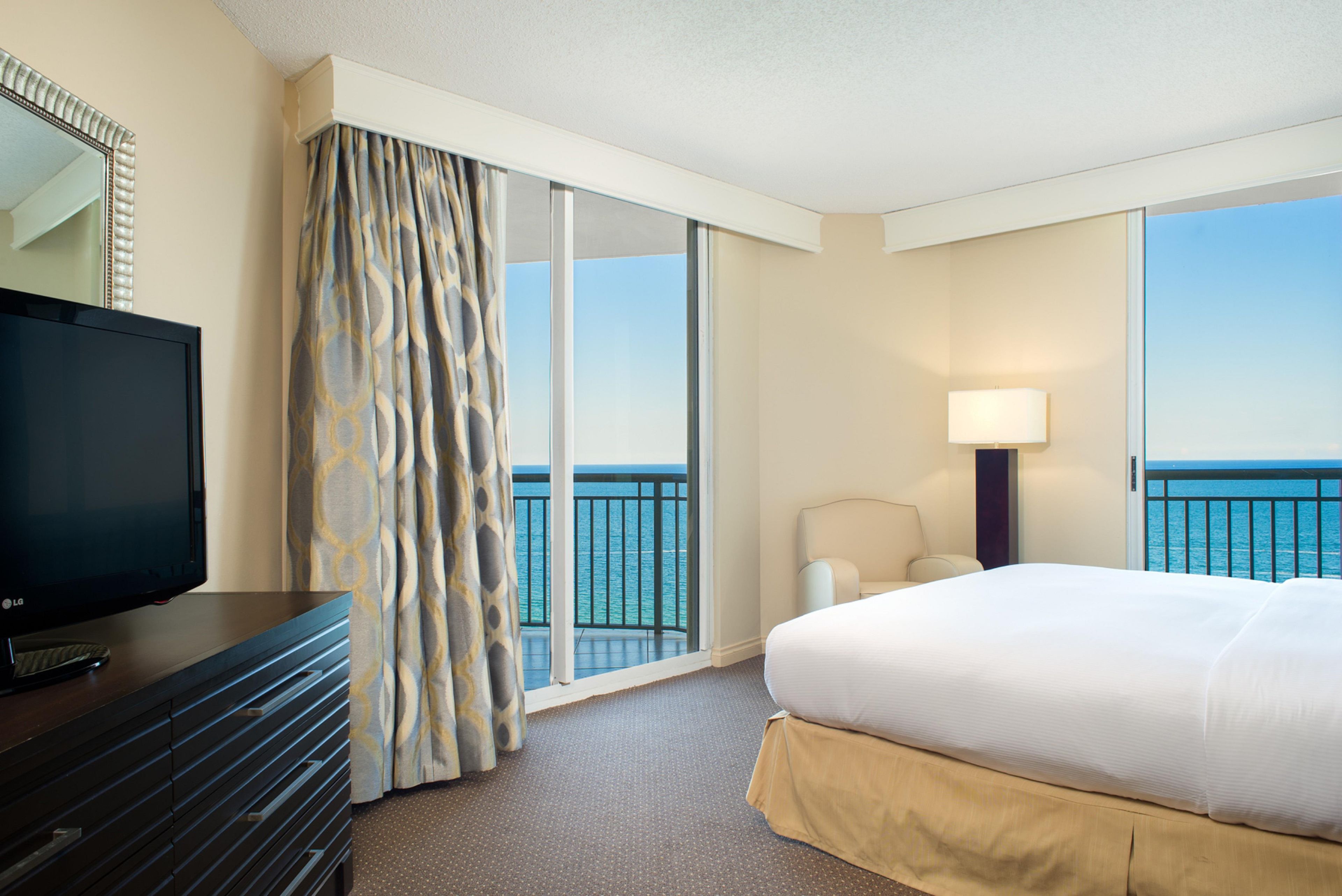 A hotel room with a bed, a TV, a chair, a lamp, and a view of the sea.