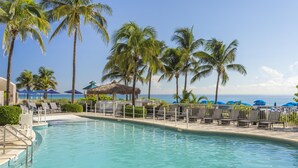A pool area with lounge chairs and umbrellas, surrounded by palm trees and a view of the ocean.