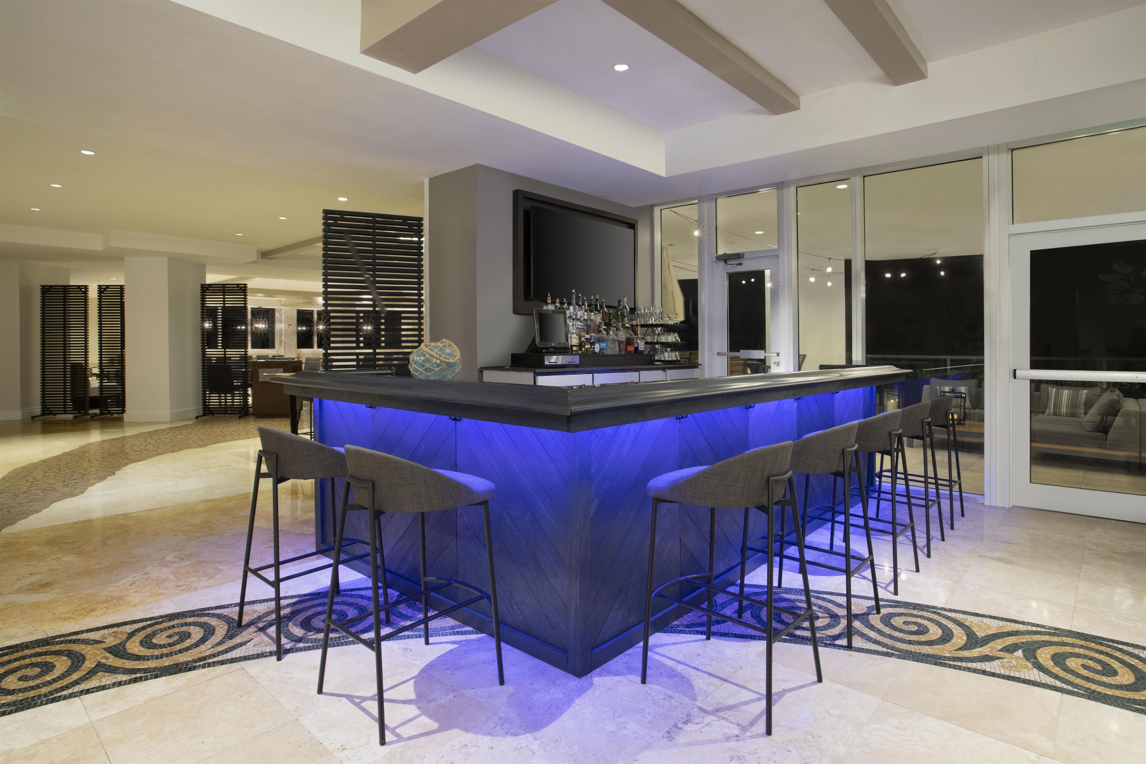A modern bar area with a blue-lit bar counter, multiple bar stools, and a television mounted above the counter.