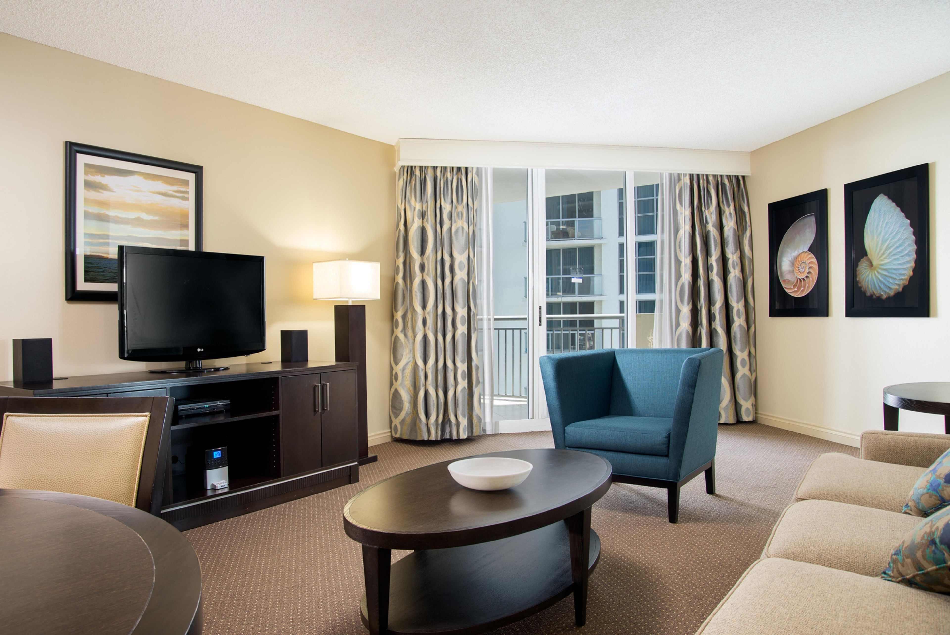 Quarto de hotel com televisão de tela plana, sofá, cadeira, mesa pequena e varanda com cortinas.