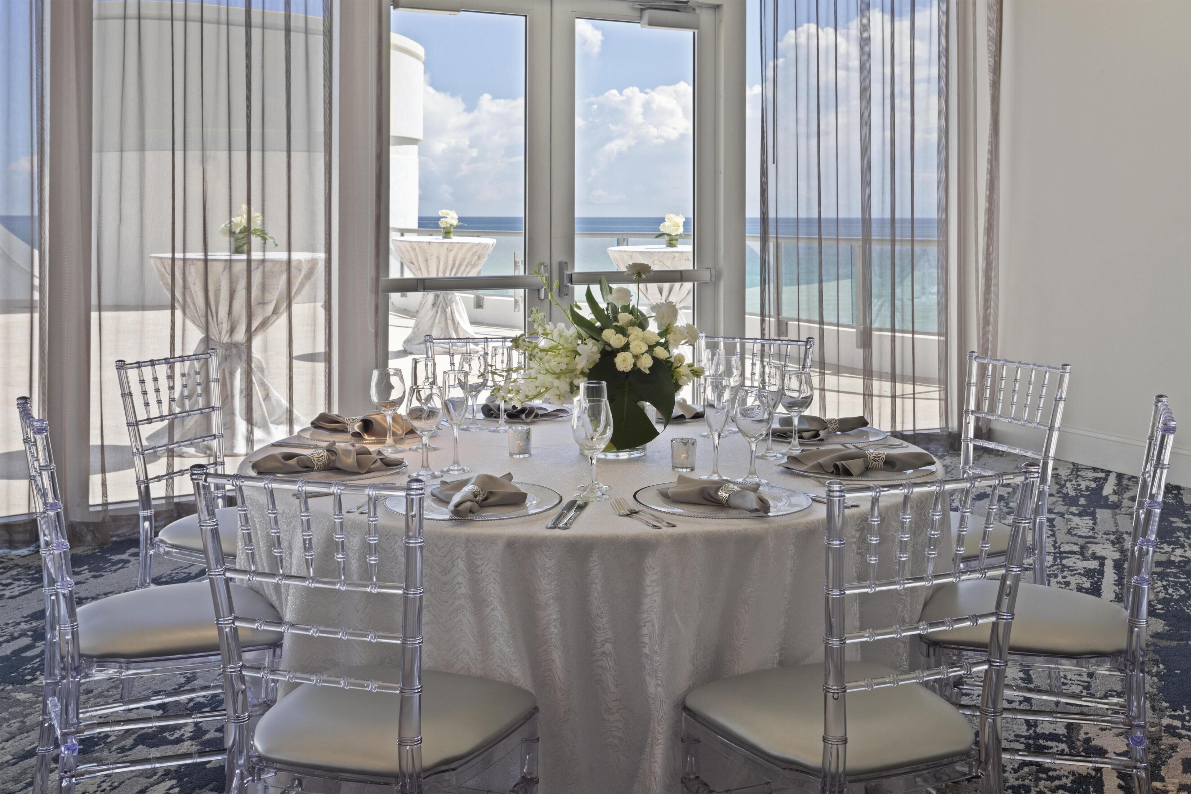 A round table with clear chairs, a white tablecloth, and a vase of white flowers.