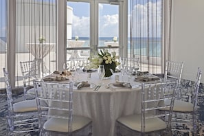 A round table set for a meal with clear chairs, a vase of flowers, and a view of the sea through large windows. - DoubleTree Resort & Spa by Hilton Ocean Point-N. Miami Beach (Sunny Isles Beach)