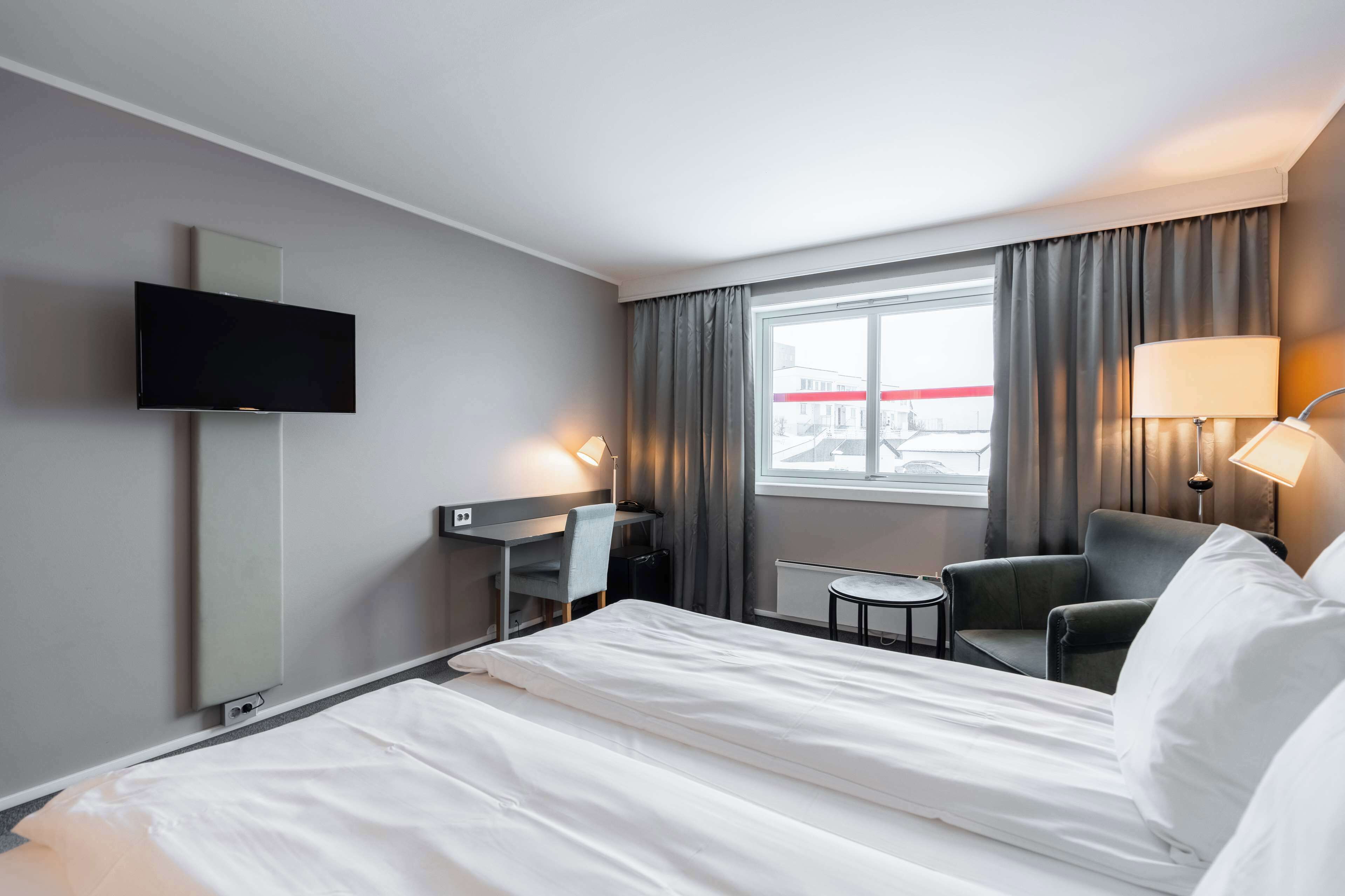 Ein Hotelzimmer mit Bett, Schreibtisch, Stuhl, Fernseher, Lampe und einem Fenster mit Vorhängen.