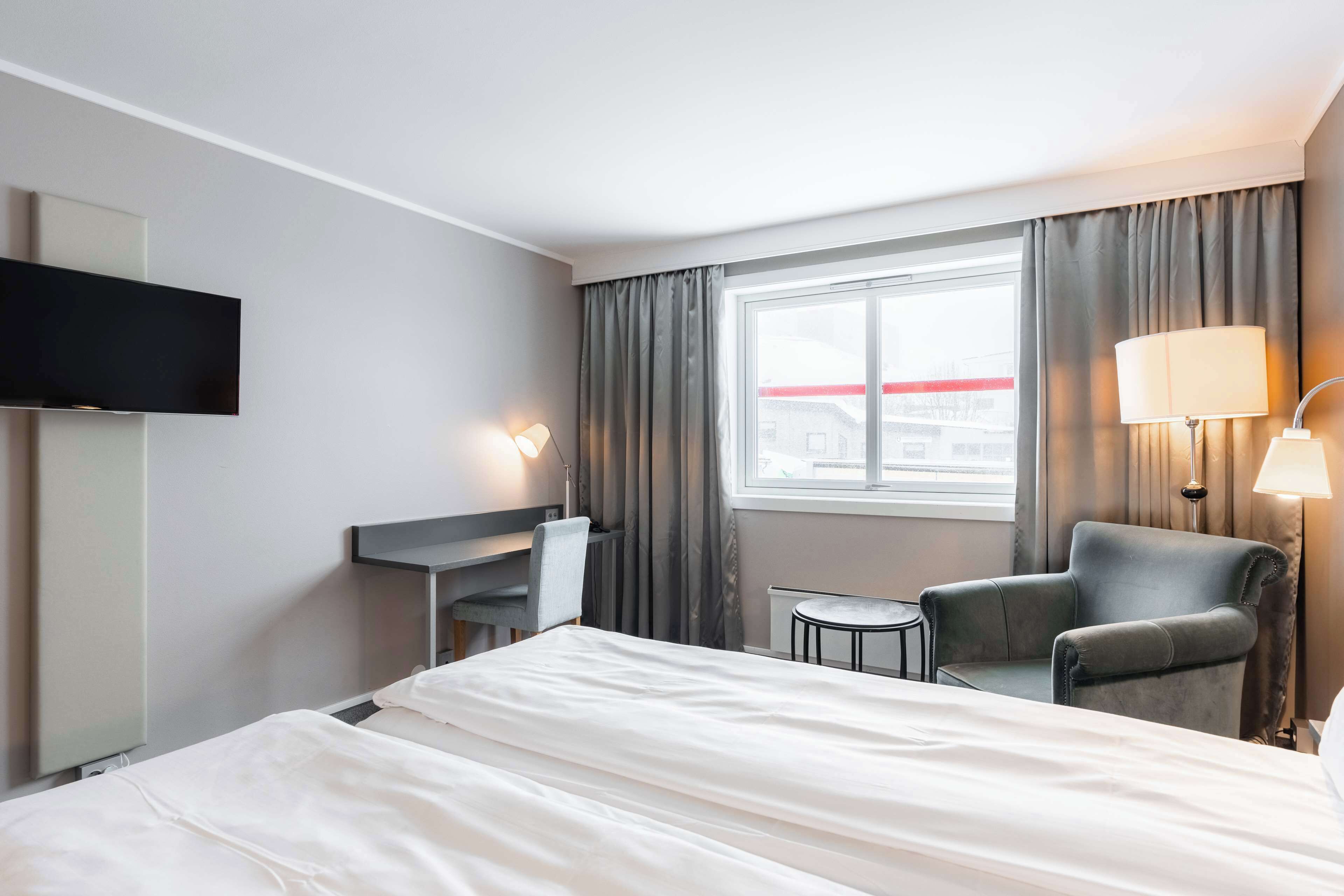 Ein Hotelzimmer mit Bett, Schreibtisch, Sessel, Fernseher und einem Fenster mit Vorhängen.